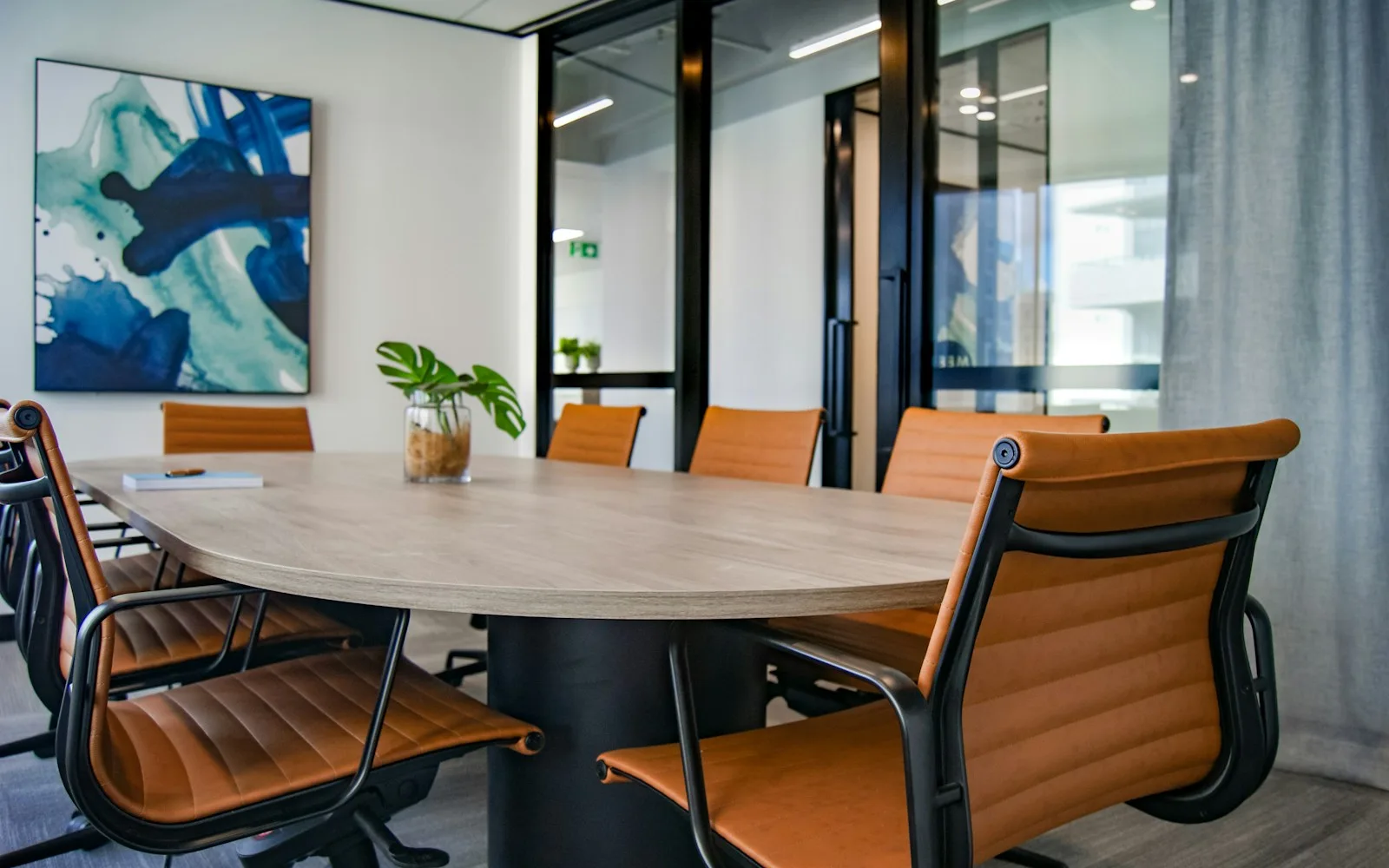 A boardroom with a curved wooden table, tan leather chairs and a small plant, glass-walled offices visible beyond.