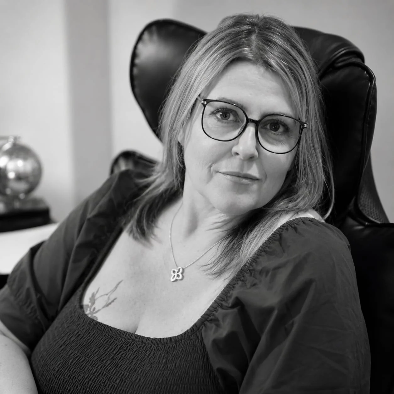 Black and white portrait of Caroline Reed seated in a leather chair, wearing glasses and smiling warmly.
